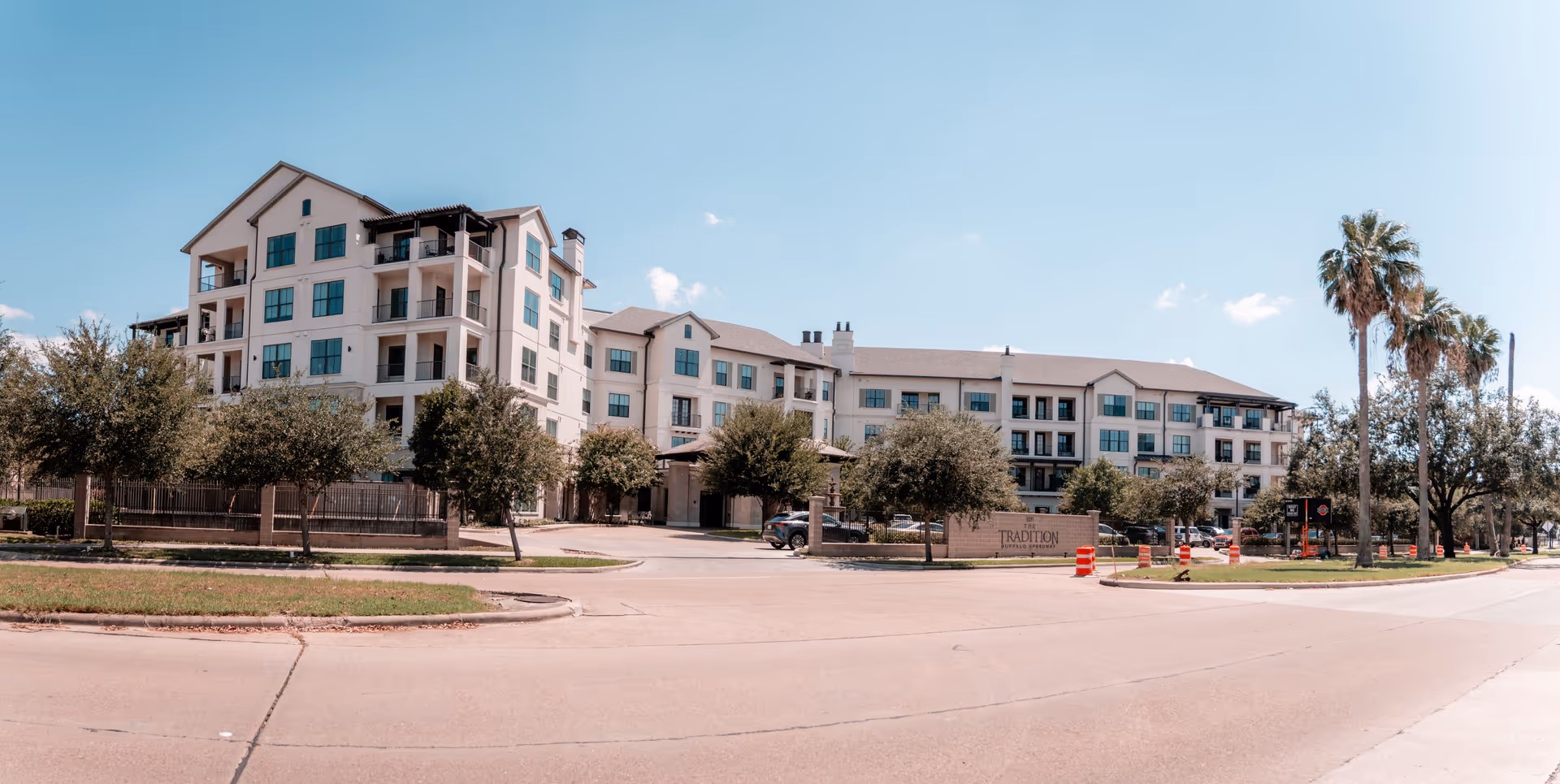 Exterior view of The Tradition–Buffalo Speedway, a multi-story senior living facility with balconies, surrounded by trees and palm trees under a clear blue sky.