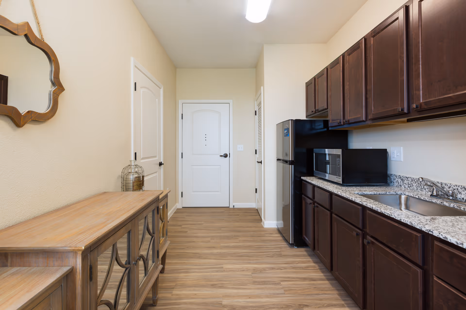 A narrow kitchen area with dark wood cabinets, a granite countertop, a stainless steel sink, a microwave, and a refrigerator. On the left side, there is a wooden sideboard with glass doors and a decorative mirror hanging above it. The floor is wood, and there are two white doors at the end of the room.