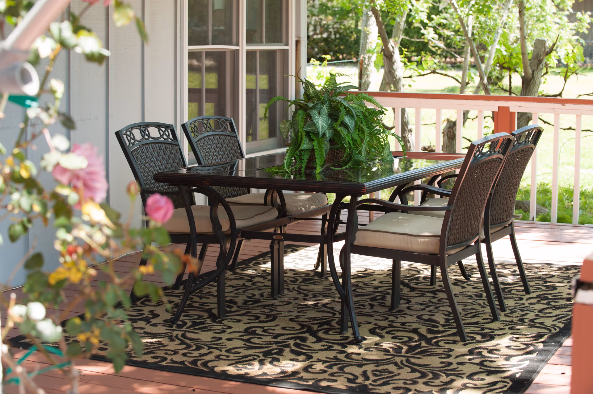 Outdoor patio area with a glass-top table surrounded by four cushioned metal chairs, a decorative patterned rug underneath, and a green potted plant centerpiece on the table. The patio is adjacent to a building with large windows and overlooks a grassy yard with trees.
