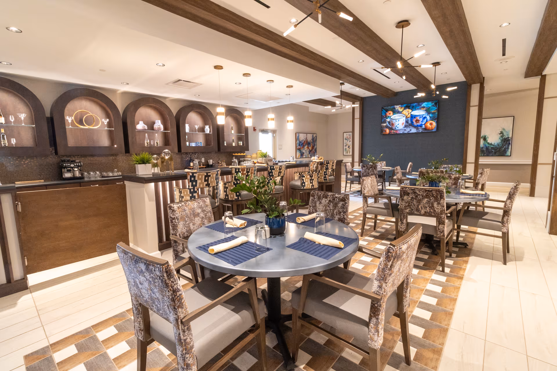 Bright, modern dining room with round tables set with napkins, upholstered chairs, a service counter/bar, and decorative lighting.