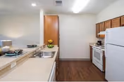 A clean and simple kitchen area with white countertops and wooden cabinets. The kitchen features a white refrigerator, white stove with oven, and a double sink. There is a small dining counter with place settings and a decorative plant. The floor is wood-style laminate, and the walls are painted white.