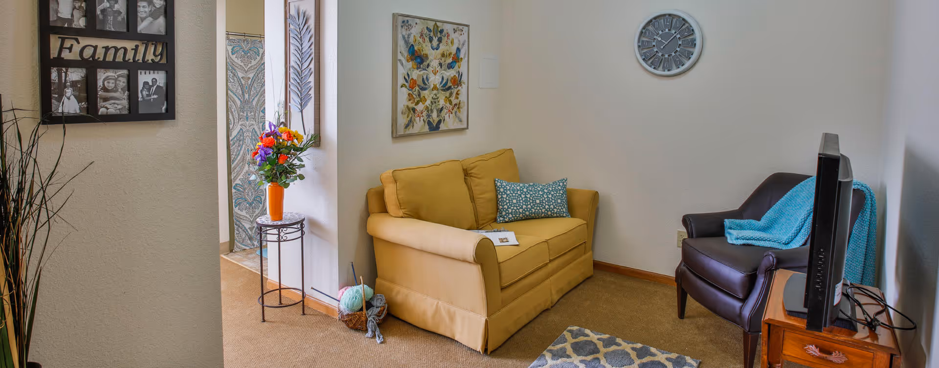 A cozy living room area with a yellow loveseat adorned with a blue patterned pillow, a dark brown armchair with a blue throw blanket, a wooden side table holding a flat-screen TV, and a wall clock above. There is a colorful floral painting on the wall and a small table with a vase of flowers near the entrance. A photo collage frame labeled 'Family' is visible on the left wall.