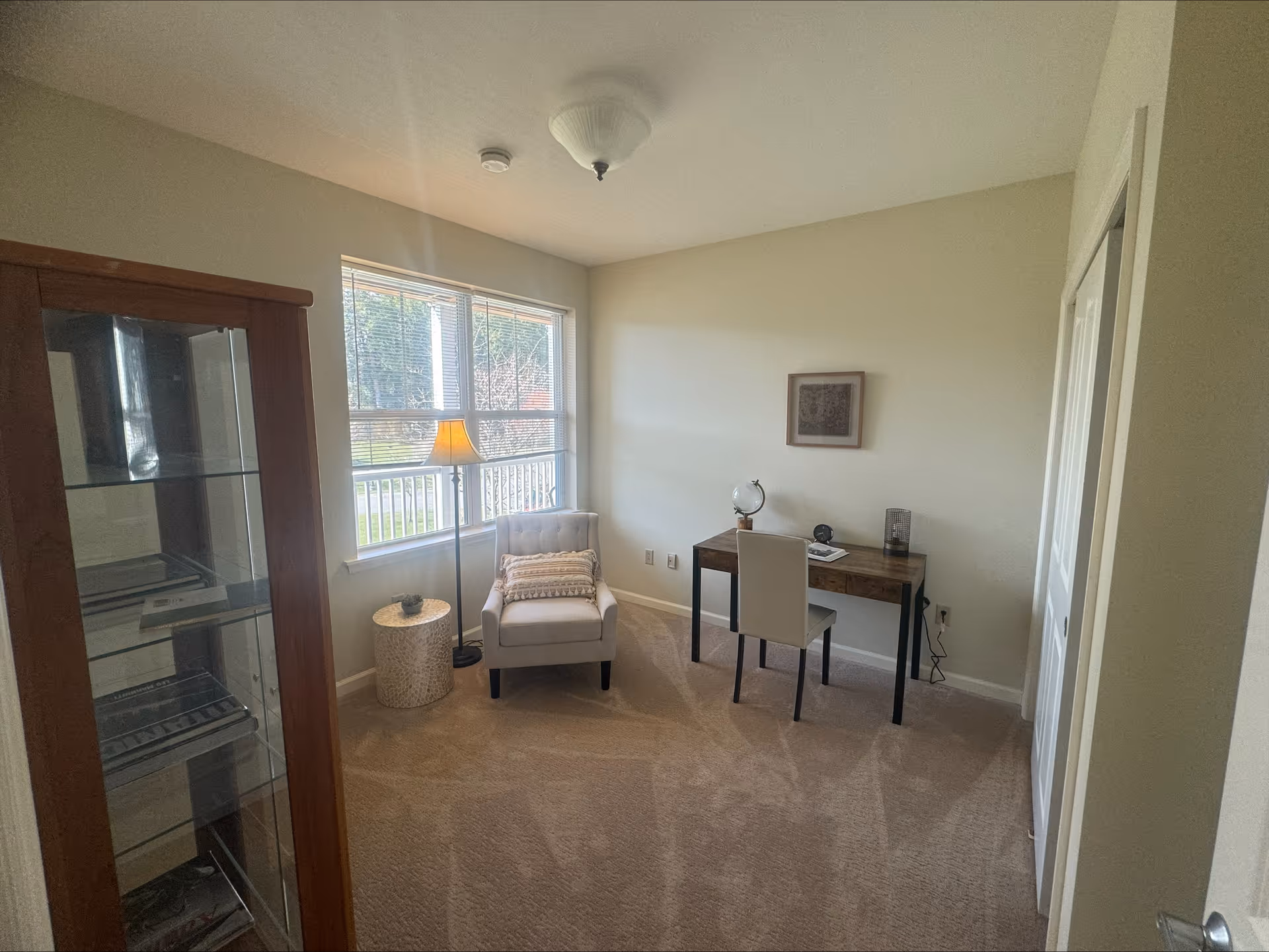 Small bright room with a desk and chair, an upholstered armchair and a floor lamp beside a window.