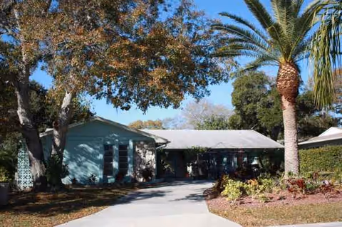 Single-story teal facility front with driveway, palm trees, and landscaped yard against blue sky.