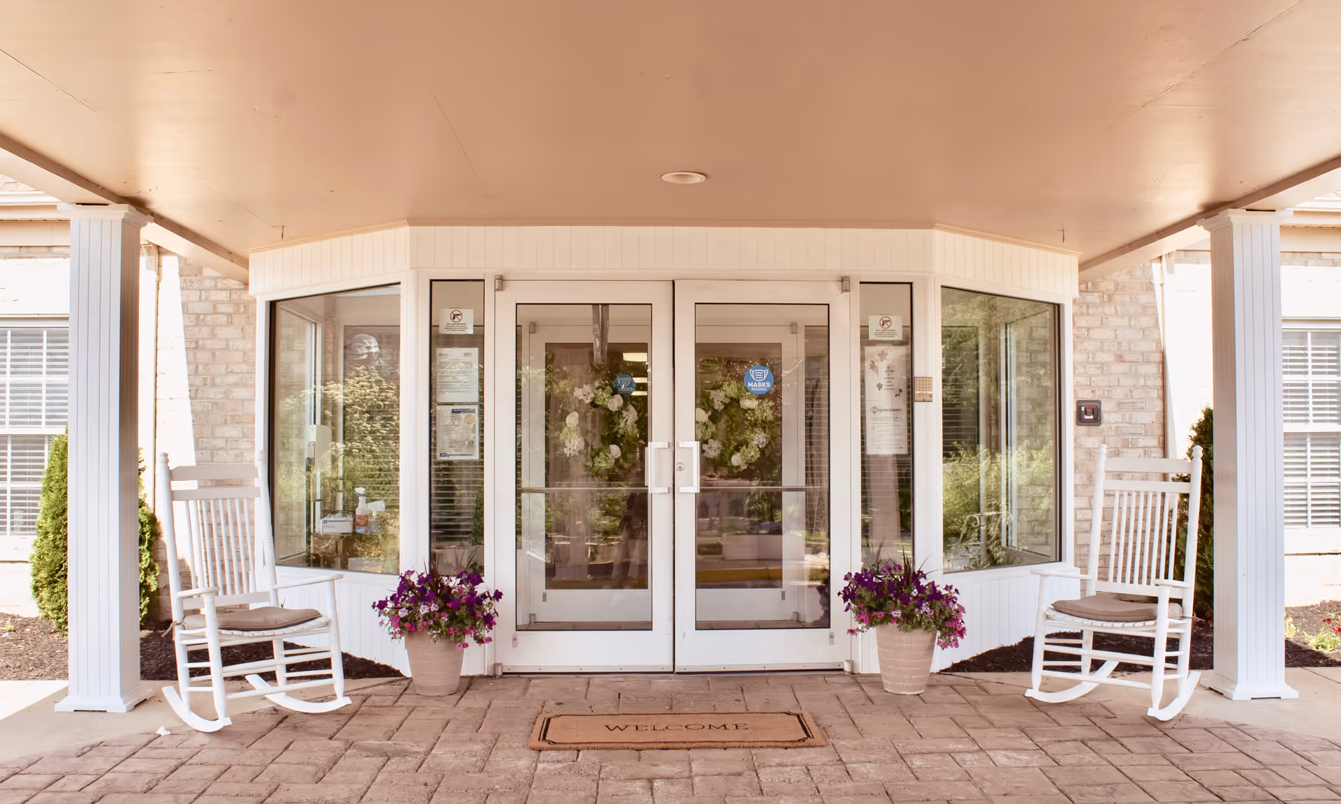 Entrance to The Maynard facility featuring double glass doors with a welcome mat in front. Two white rocking chairs with cushions are placed on either side of the entrance, accompanied by potted purple flowers. The area is covered by a roof supported by white columns.