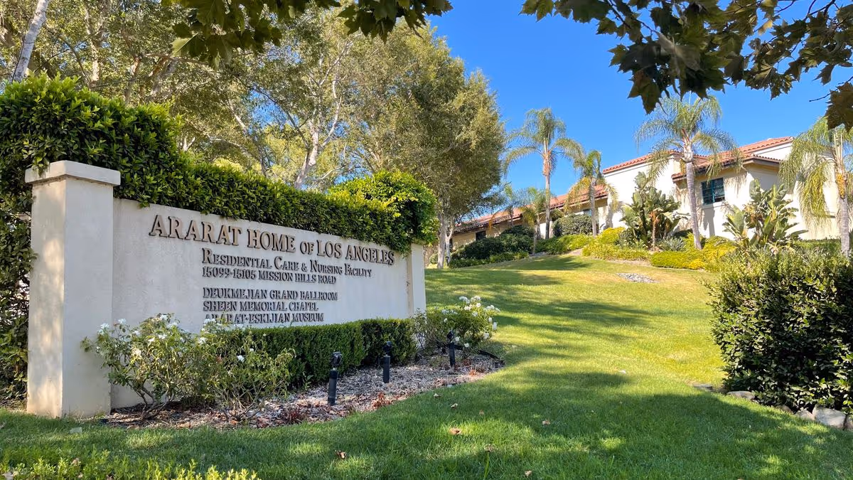 Entrance sign reading "Ararat Home of Los Angeles" on a landscaped lawn with the facility buildings, palm trees, and blue sky in the background.