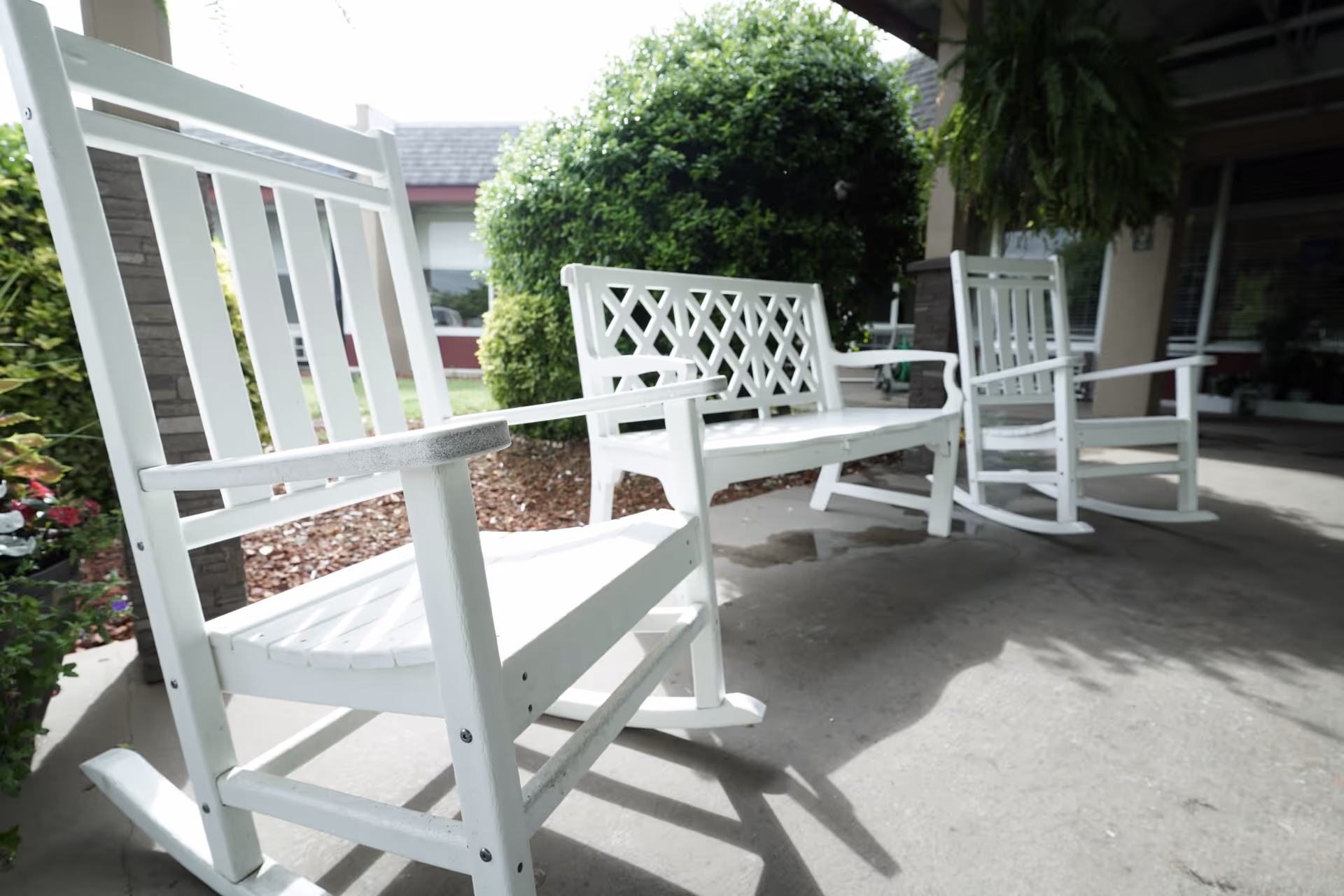 White wooden rocking chairs and a bench sit on a covered front porch with shrubs in the background.