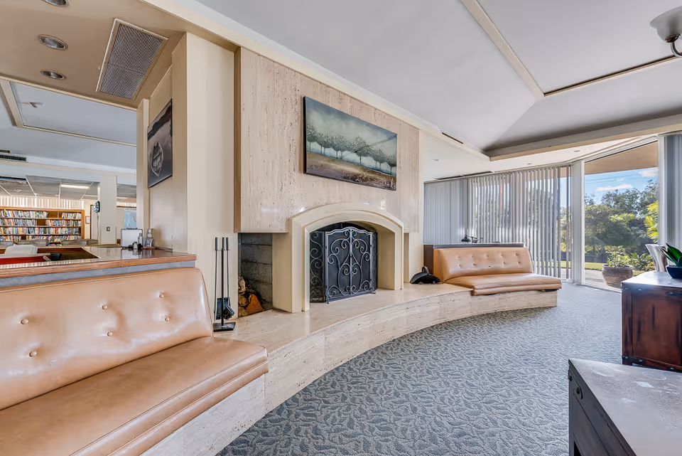A spacious living room area with a curved beige stone fireplace in the center. Two tan leather cushioned benches are built into the curved stone hearth on either side of the fireplace. A painting of trees hangs above the fireplace. Large windows with vertical blinds allow natural light to fill the room and provide a view of greenery outside. In the background, there is a glimpse of a library with bookshelves and seating.