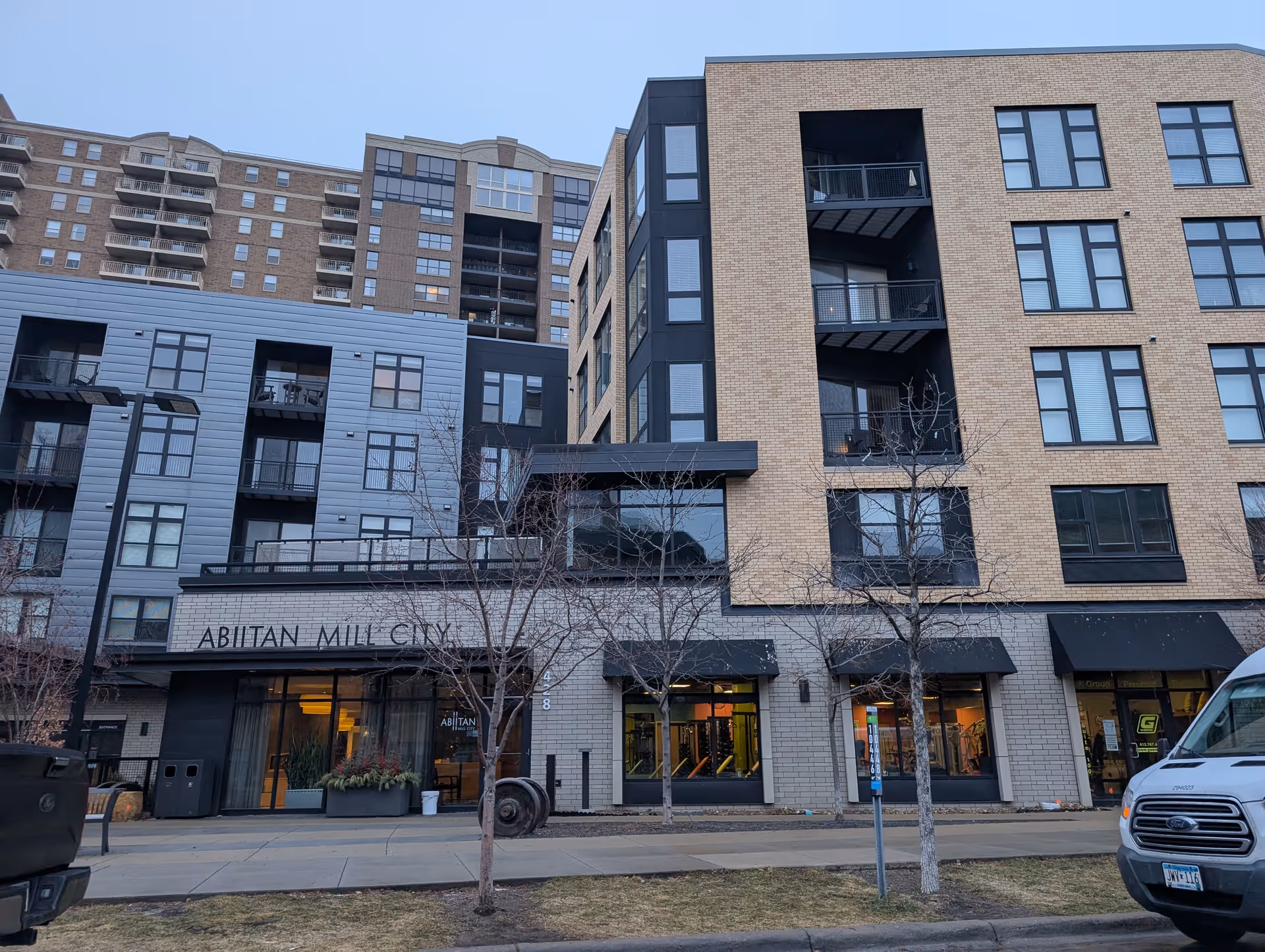 Exterior view of Abiitan Mill City building showing a modern multi-story structure with large windows and balconies. There are leafless trees and parked vehicles in front of the building.