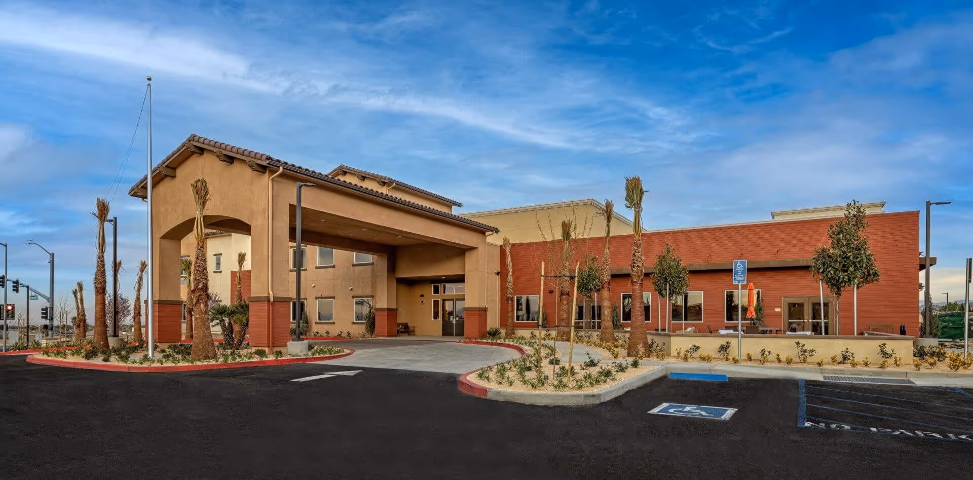 Exterior view of Palm Vista Senior Living facility showing the entrance with a covered drop-off area, palm trees, and a parking lot with handicapped parking spaces under a partly cloudy blue sky.