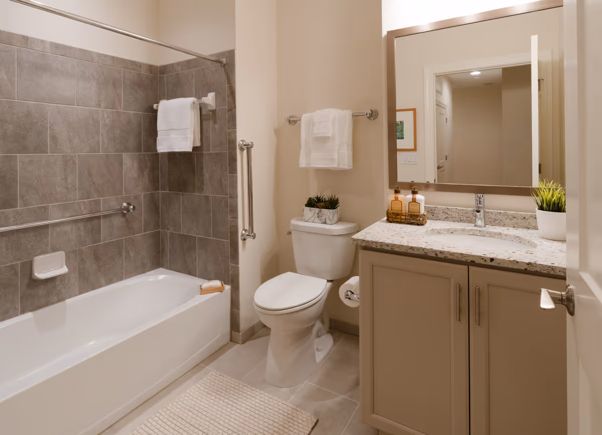 A clean and modern bathroom featuring a bathtub with gray tiled walls and a white towel hanging on a rod above it. There is a white toilet next to the bathtub with a towel rack above it holding a folded white towel. A vanity with a granite countertop, a sink, two soap dispensers, and a small potted plant is visible. A large mirror hangs above the vanity, reflecting part of the hallway outside the bathroom.