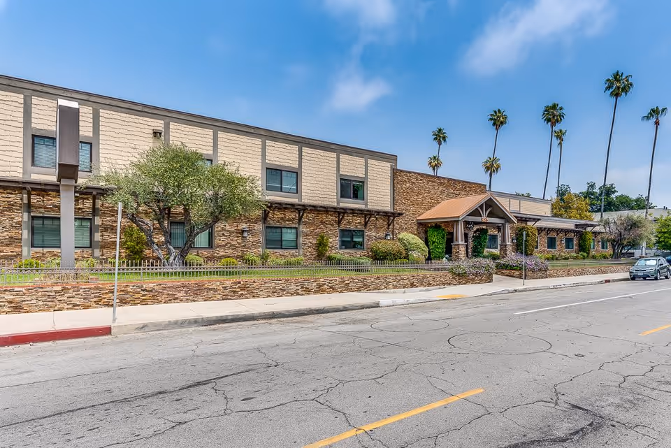 Exterior view of a senior living facility building with stone and beige siding, a covered entrance, palm trees in the background, and a car parked on the street in front.
