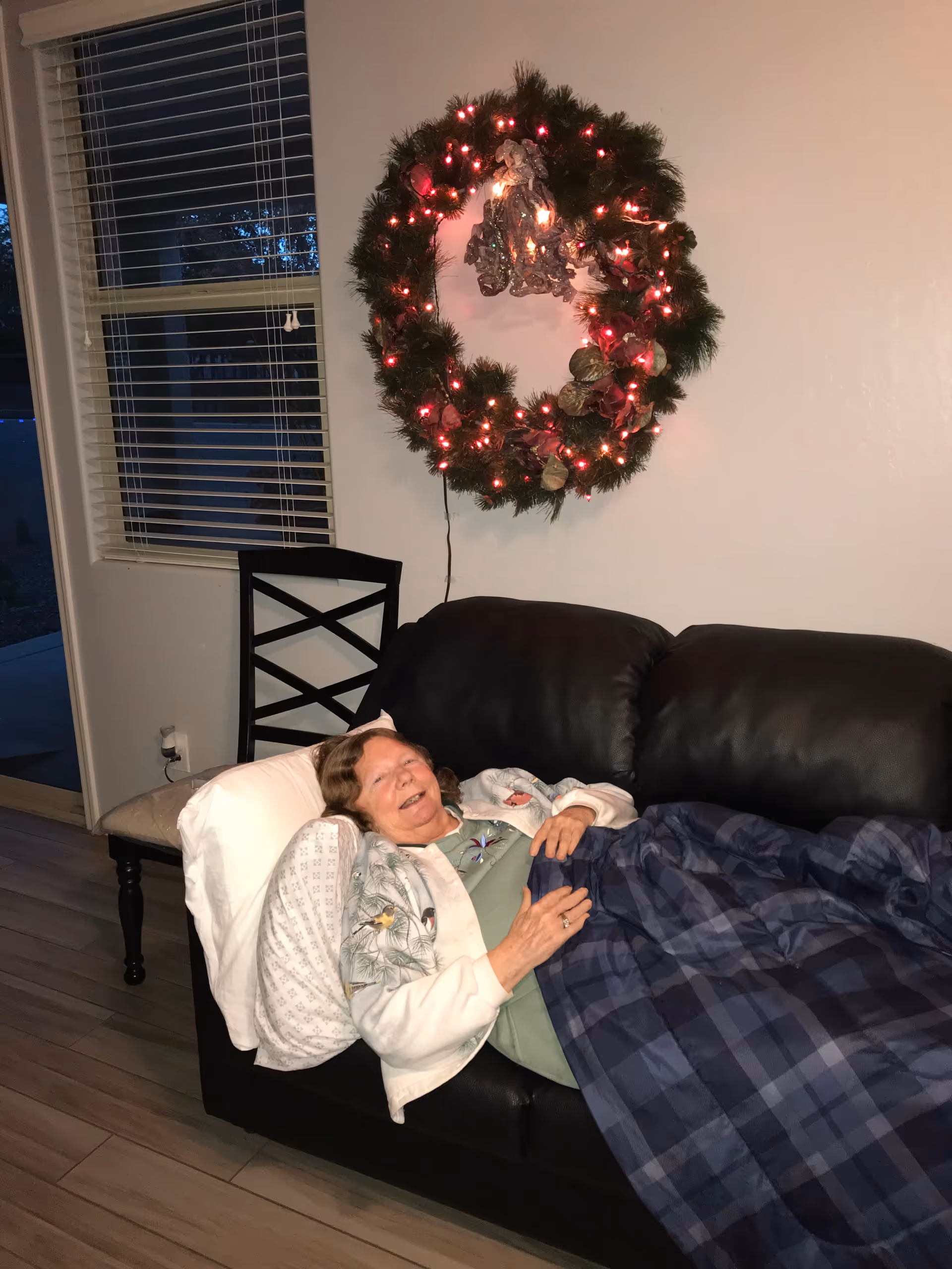 An elderly woman lies on a black leather couch covered by a plaid blanket and pillow, smiling beneath a lit holiday wreath on the wall.