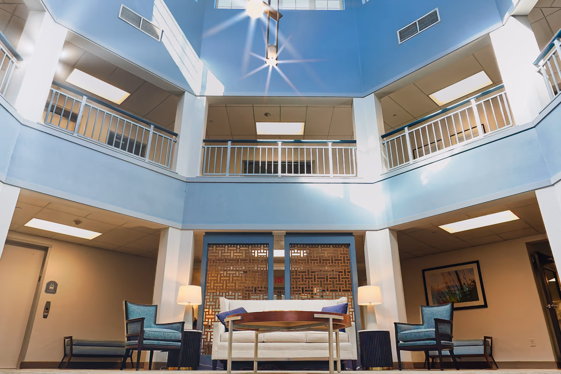 Interior view of a spacious, well-lit common area in a senior living facility with a high ceiling and skylights. The room features a white sofa with blue cushions, two blue armchairs, a wooden coffee table, two lamps, and decorative wooden panels behind the sofa. There are railings on the upper level surrounding the open space.