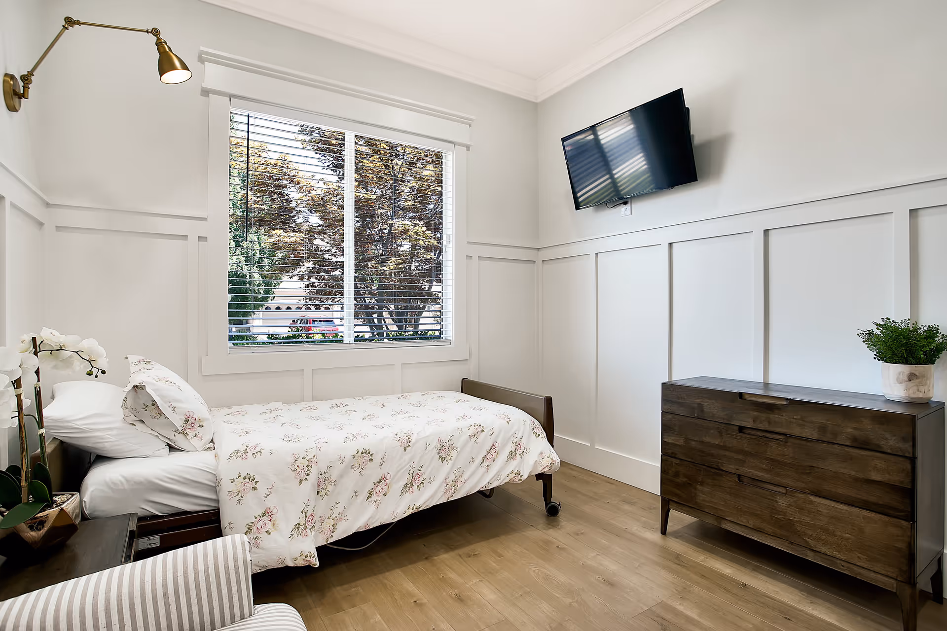 A bright and clean bedroom with a single bed covered in floral bedding, a wooden dresser with a potted plant on top, a wall-mounted flat-screen TV, a large window with white blinds showing trees outside, and a striped armchair partially visible in the foreground.