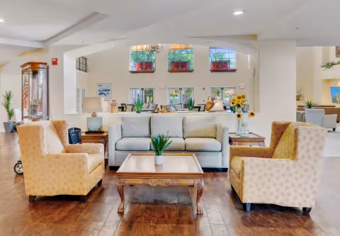 Bright senior living lounge with a sofa, four armchairs and a wooden coffee table in an open lobby area with windows and decor.