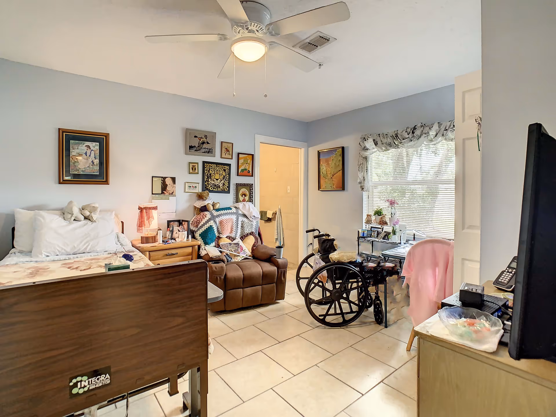 A senior living facility bedroom with a hospital-style bed, a brown recliner chair covered with a colorful crocheted blanket, a wheelchair, and a window with blinds and a valance. The room has light blue walls decorated with various framed pictures and a ceiling fan with a light. There is a small wooden nightstand with a lamp and personal items, and a door leading to a bathroom.