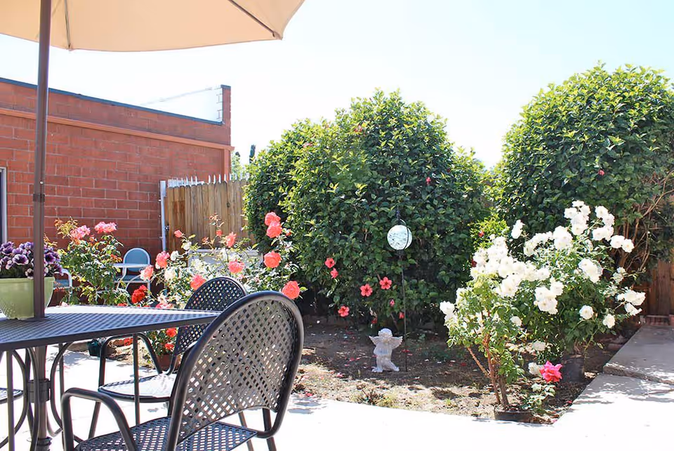 Outdoor patio area with a black metal table and chairs under a large beige umbrella. Surrounding the patio are various flowering bushes including pink and white roses, with a small angel statue and a decorative hanging ornament among the plants. A brick wall and wooden fence are visible in the background under a clear sky.