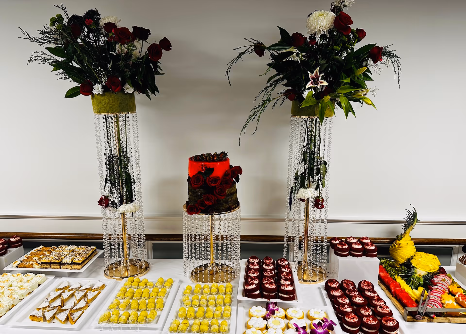 A dessert buffet table featuring an assortment of pastries, cupcakes, and fruit arrangements. The table is decorated with two tall floral arrangements with red roses and white flowers in crystal bead stands. There is a red and brown cake in the center, surrounded by various trays of yellow and red desserts and a pineapple fruit carving on the right side.
