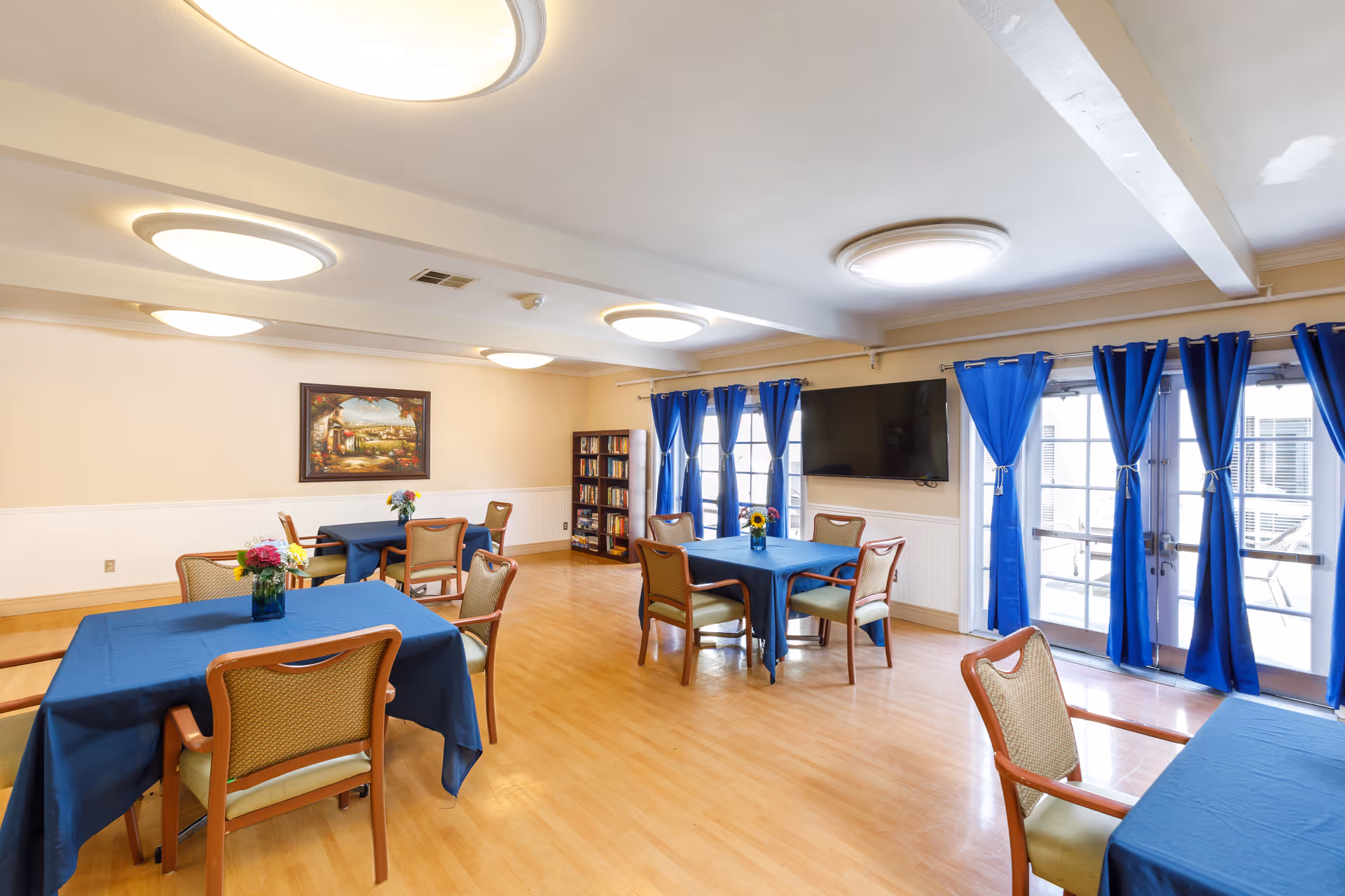 A bright and spacious common area with several tables covered in blue tablecloths, each surrounded by chairs. The room has wooden flooring, large windows with blue curtains, a wall-mounted TV, a bookshelf, and a framed painting on the wall.