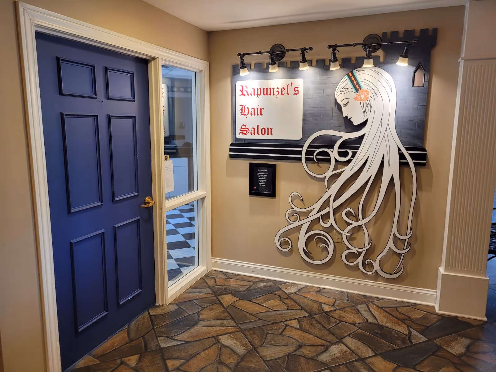 Interior view of a hallway corner with a blue door on the left and a decorative wall sign on the right that reads 'Rapunzel's Hair Salon' with an artistic depiction of a woman with long flowing hair. The floor has a stone tile pattern and the walls are beige.