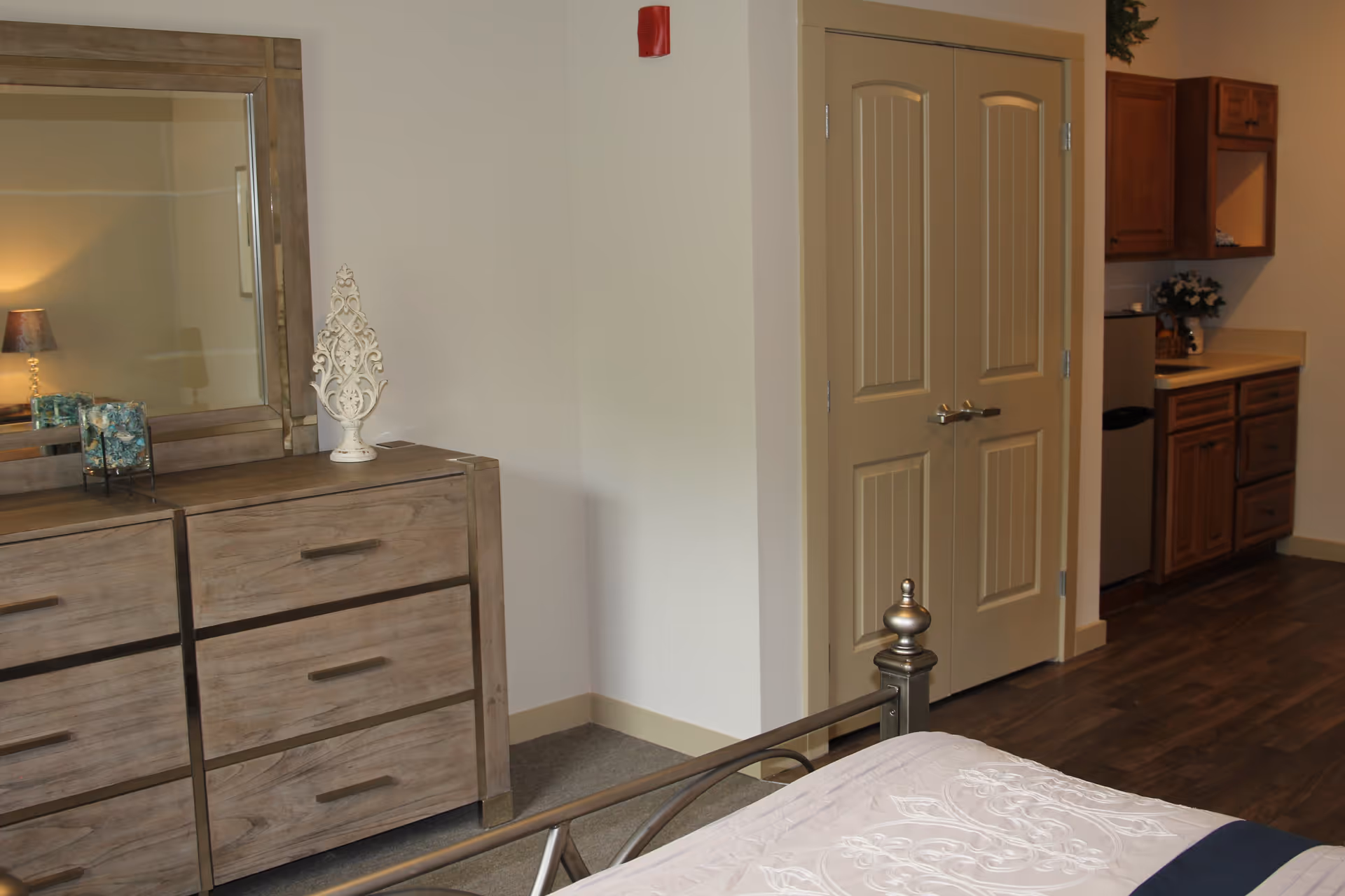 A bedroom area in a senior living facility featuring a wooden dresser with a large mirror, decorative items on top, a metal bed frame with a white and blue patterned bedspread, a double door closet, and a small kitchenette with wooden cabinets and a mini refrigerator.