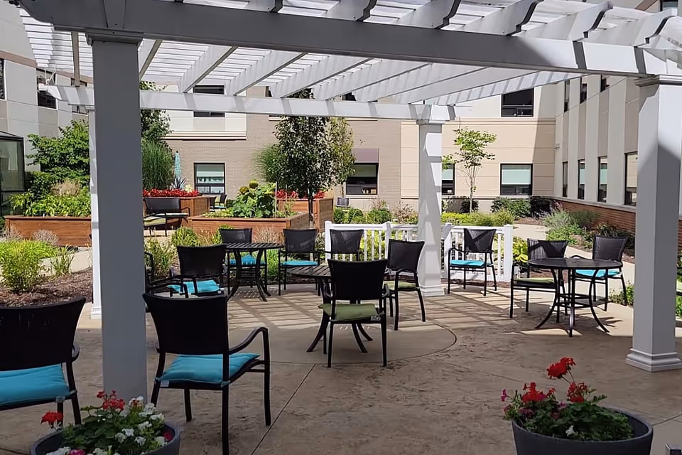 Outdoor patio area with black metal chairs featuring blue and green cushions, black round tables, and a white pergola overhead. Surrounding the patio are raised garden beds with various plants and flowers, and the background shows the exterior walls and windows of a building.