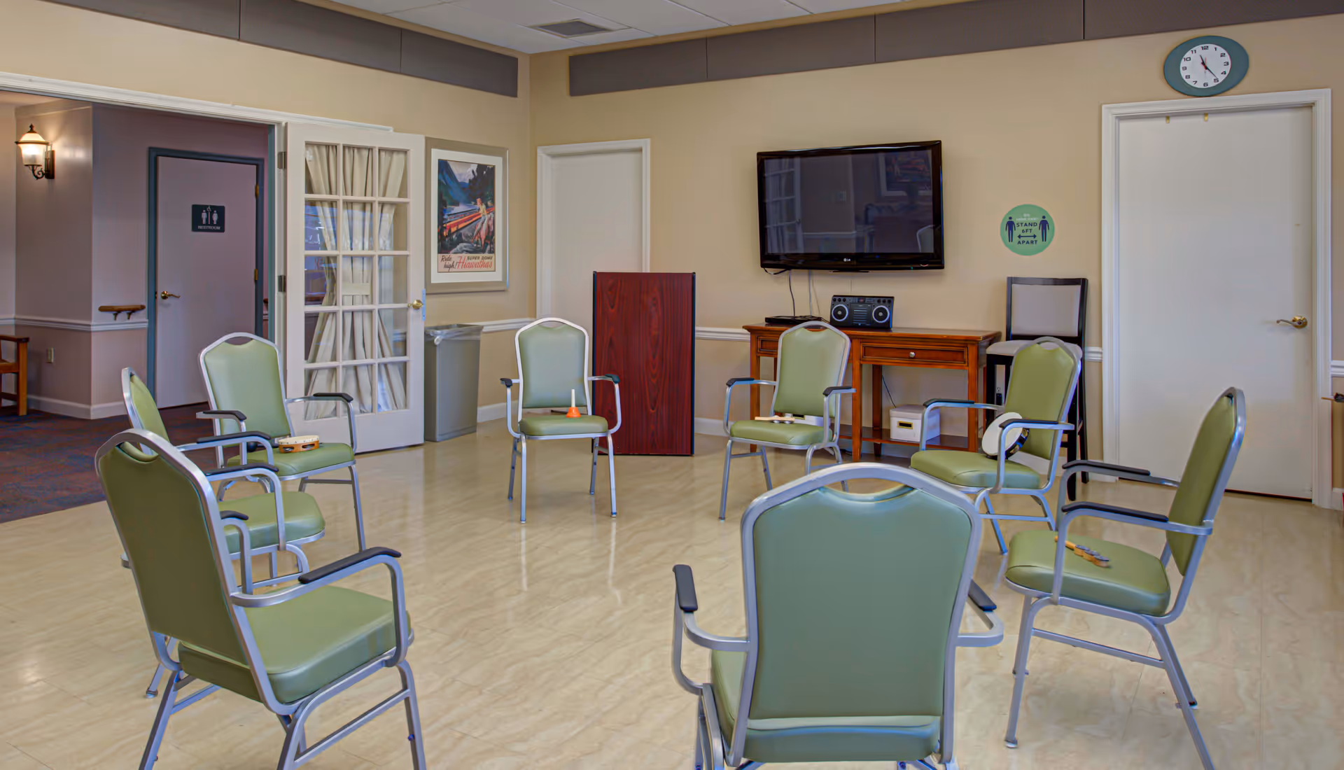 A room with green cushioned chairs arranged in a circle on a light-colored floor. The room has beige walls with white trim, a wall-mounted TV above a wooden table with a stereo, and a clock showing 11:55. There are two closed white doors, one with a sign indicating a restroom, and a glass door with curtains. A poster and a trash can are visible near the glass door. Some chairs have small musical instruments like a tambourine and a bell placed on them.