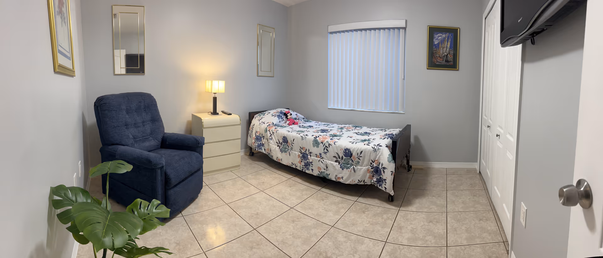 A tidy bedroom featuring a single bed with a floral comforter, a blue recliner, a nightstand with a lamp, a wall-mounted TV, closet doors and tiled floor.