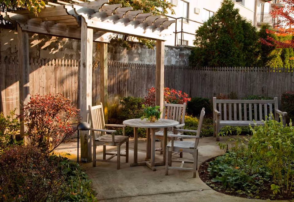 Sunlit courtyard with a wooden pergola, round outdoor table and chairs, benches, and surrounding plants and a wooden fence.