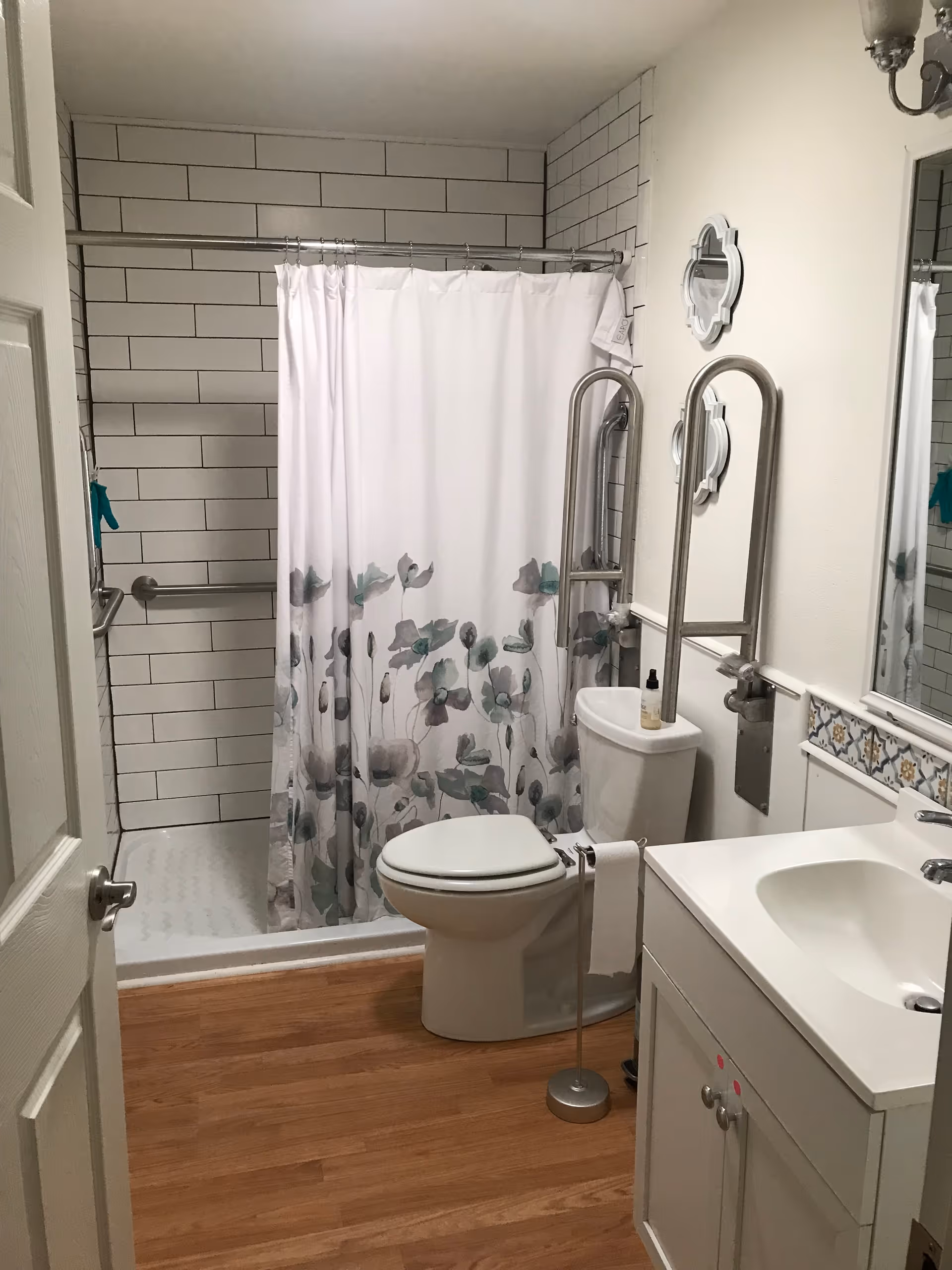 A clean and accessible bathroom with a white toilet, a white sink with a cabinet, a shower area with white subway tiles and a floral shower curtain, and safety grab bars installed near the toilet and shower. The floor is wooden, and there is a small towel holder with a white towel next to the toilet.