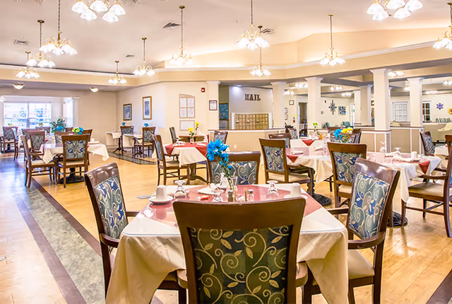 A bright and spacious dining room with multiple tables covered in white tablecloths and set with cups, glasses, and napkins. The chairs have patterned upholstery, and there are blue flower centerpieces on the tables. The room features warm lighting with multiple chandeliers and large windows letting in natural light.