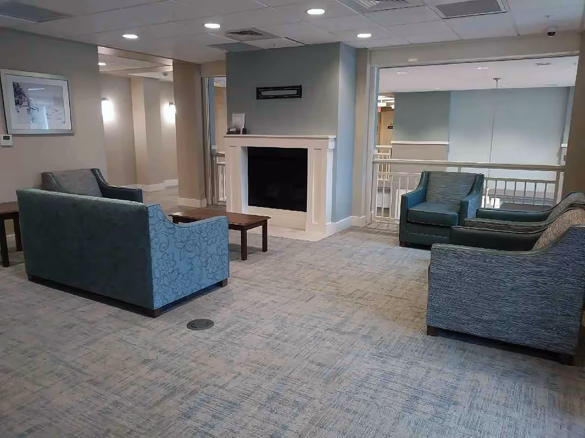 A senior living facility common area with several upholstered chairs and sofas arranged around a small wooden coffee table. The room features a light blue and beige color scheme, carpeted floor, a white fireplace, and overhead recessed lighting. There is a railing overlooking a lower level in the background.