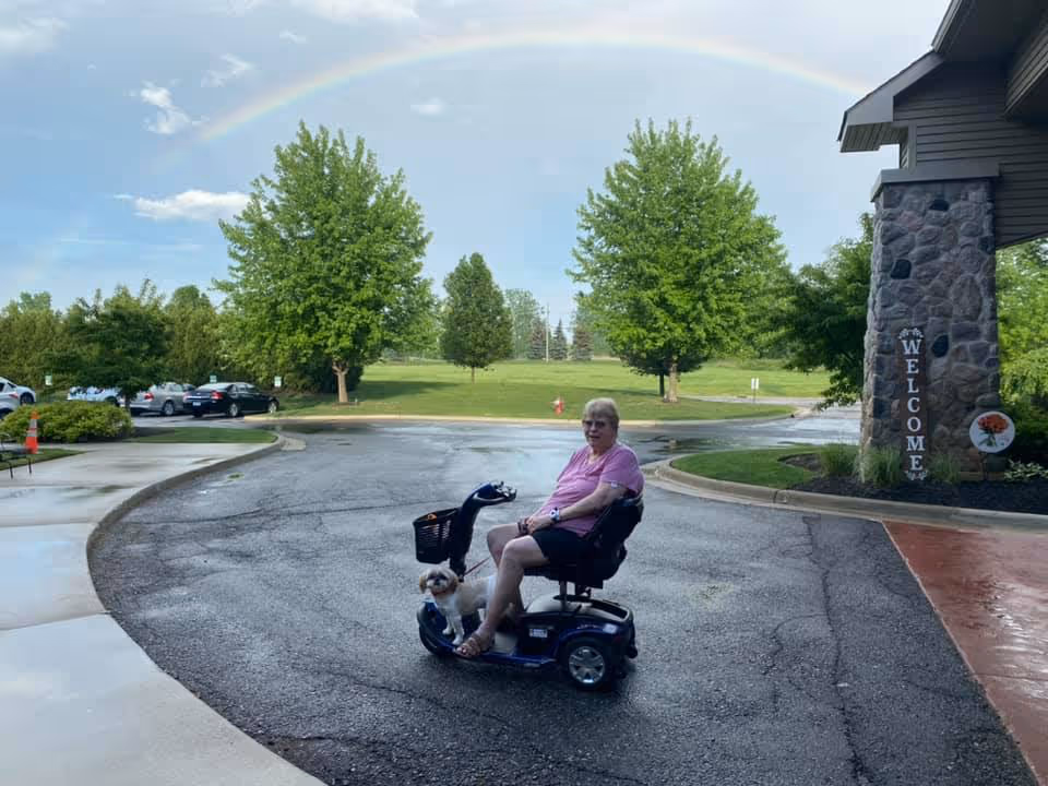 A person on a mobility scooter with a small dog in front, parked outside a building under a rainbow.