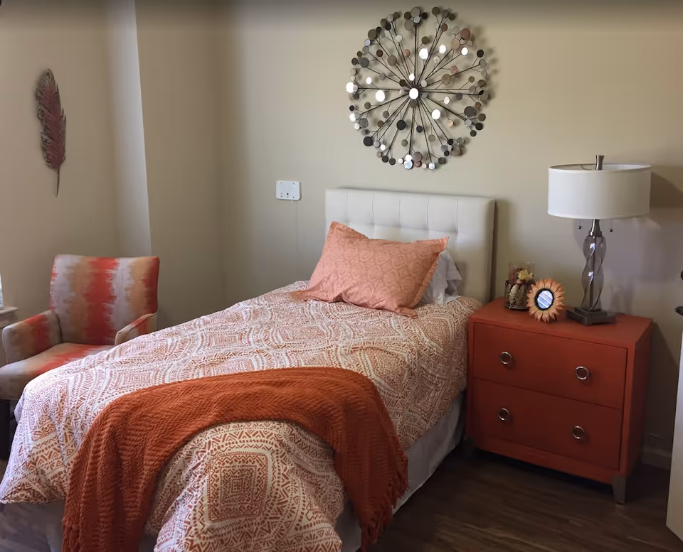 A neatly made single bed with orange patterned bedding, a matching orange nightstand and chair in a decorated bedroom.