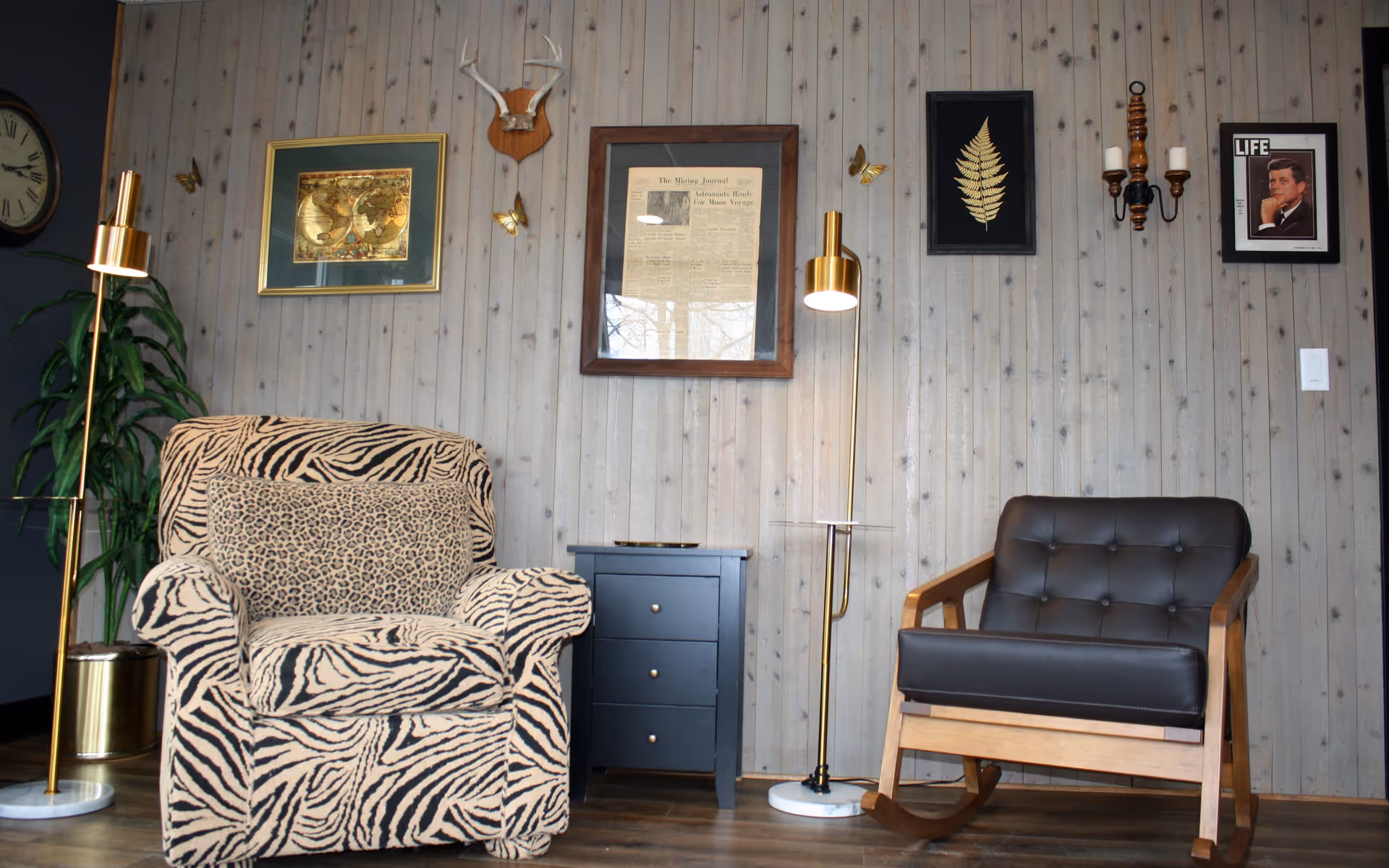 A cozy sitting area with a zebra-patterned armchair and a leopard-print pillow on the left, a black three-drawer side table in the center, and a black leather cushioned wooden rocking chair on the right. Two gold floor lamps with white marble bases flank the chairs. The wall behind is wooden paneling decorated with framed pictures, a mounted antler, and a wall sconce with two candles.