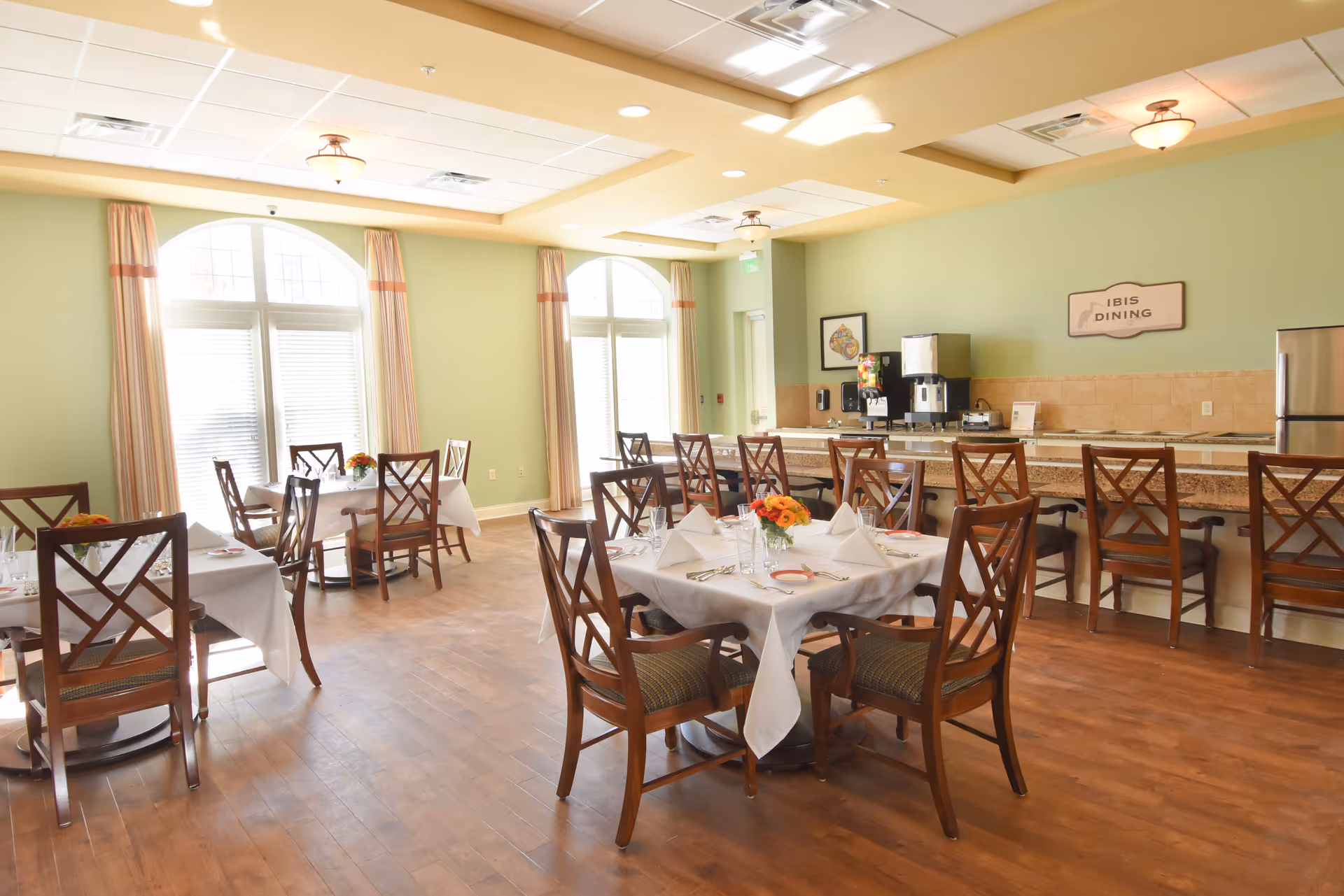 Sunny dining room with tables set for meals, wooden chairs, and a counter labeled 'IBIS DINING'.