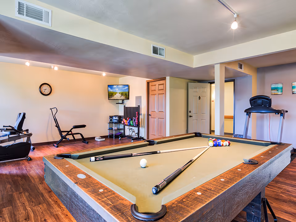A recreational room featuring a pool table with cues and balls arranged for a game, exercise equipment including a treadmill and a stationary bike, a wall-mounted TV, and a clock on the wall. The room has wooden flooring and neutral-colored walls with some artwork.