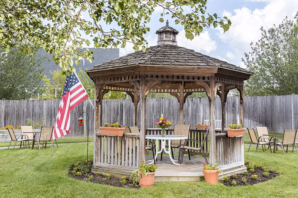 A wooden gazebo with a shingled roof situated in a grassy backyard area. The gazebo has flower pots with colorful flowers on its railing and a white round table with chairs inside. Surrounding the gazebo are additional chairs and an American flag on a pole. Trees and a wooden fence enclose the area under a partly cloudy sky.