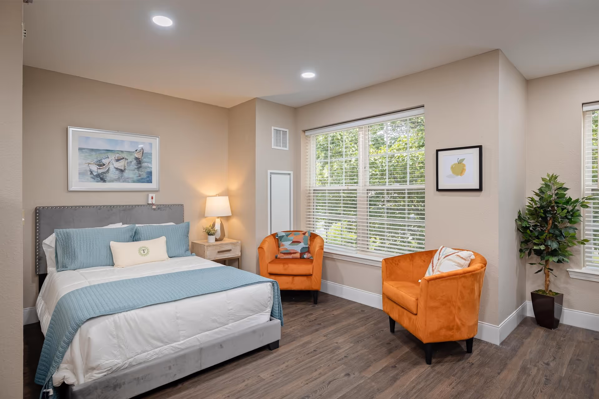 A bright and cozy bedroom featuring a neatly made bed with light blue bedding and a gray upholstered headboard. There is a wooden nightstand with a lamp and a small plant beside the bed. Two orange armchairs with decorative pillows are positioned near large windows with white blinds, allowing natural light to fill the room. A framed painting of boats on water hangs above the bed, and a framed picture of a yellow apple is on the adjacent wall. A potted plant is placed in the corner near the window.