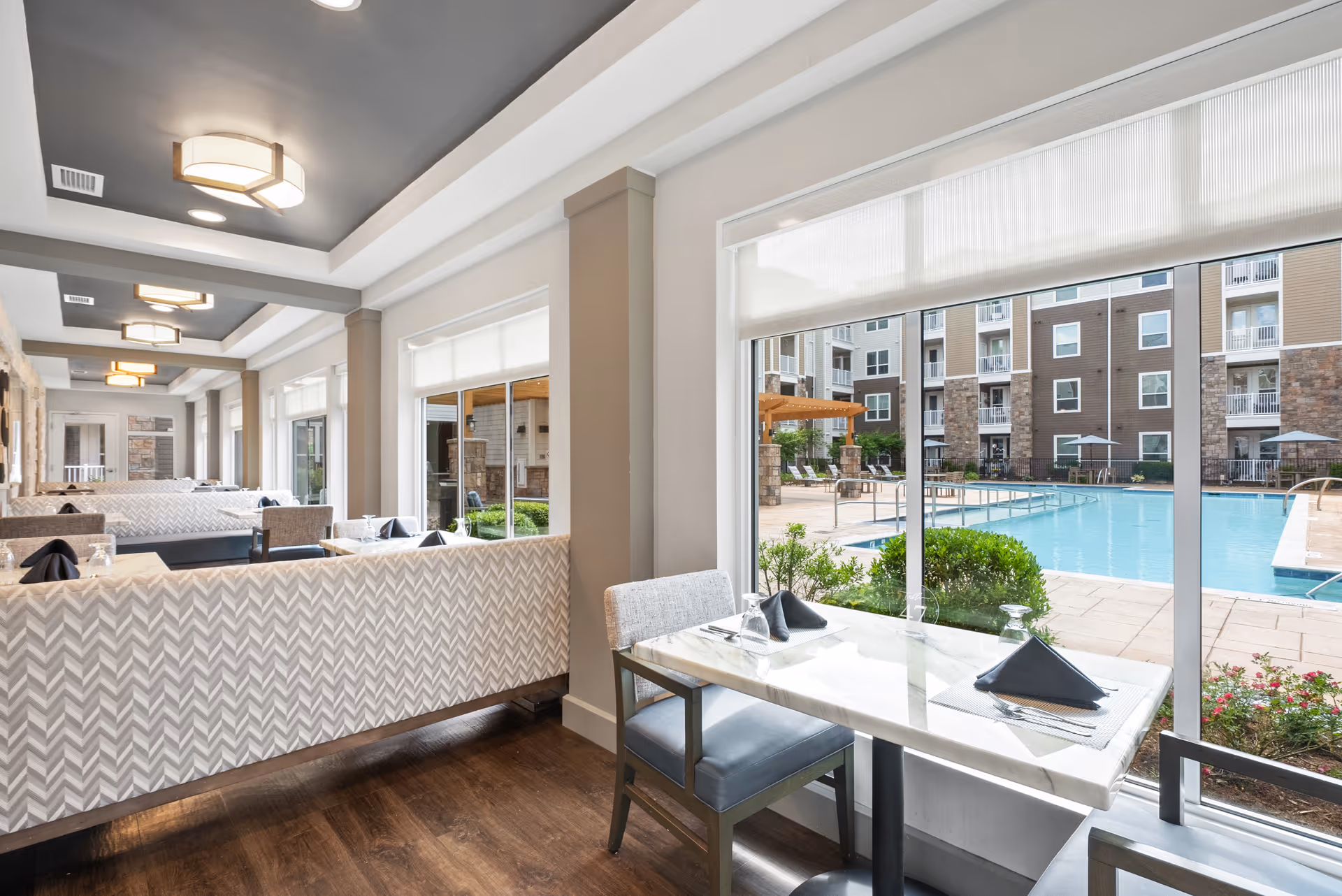 Interior dining area with tables and chairs set with napkins and glasses, large windows offering a view of an outdoor swimming pool and apartment building.