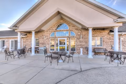 Outdoor patio area with several black metal tables and chairs arranged on a concrete surface in front of a brick building with large windows and white columns supporting a peaked roof.