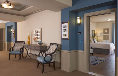 A well-lit interior corridor seating area with two upholstered chairs, a console table with decor, and an open doorway revealing a furnished bedroom.