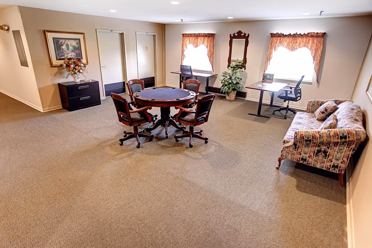 A spacious room with beige carpet and walls, featuring a round wooden table with four cushioned chairs in the center. There is a floral-patterned couch with matching pillows on the right side, two desks with office chairs near windows with floral curtains, a tall mirror on the wall, a potted plant, and a small black cabinet with a flower arrangement and framed artwork above it.
