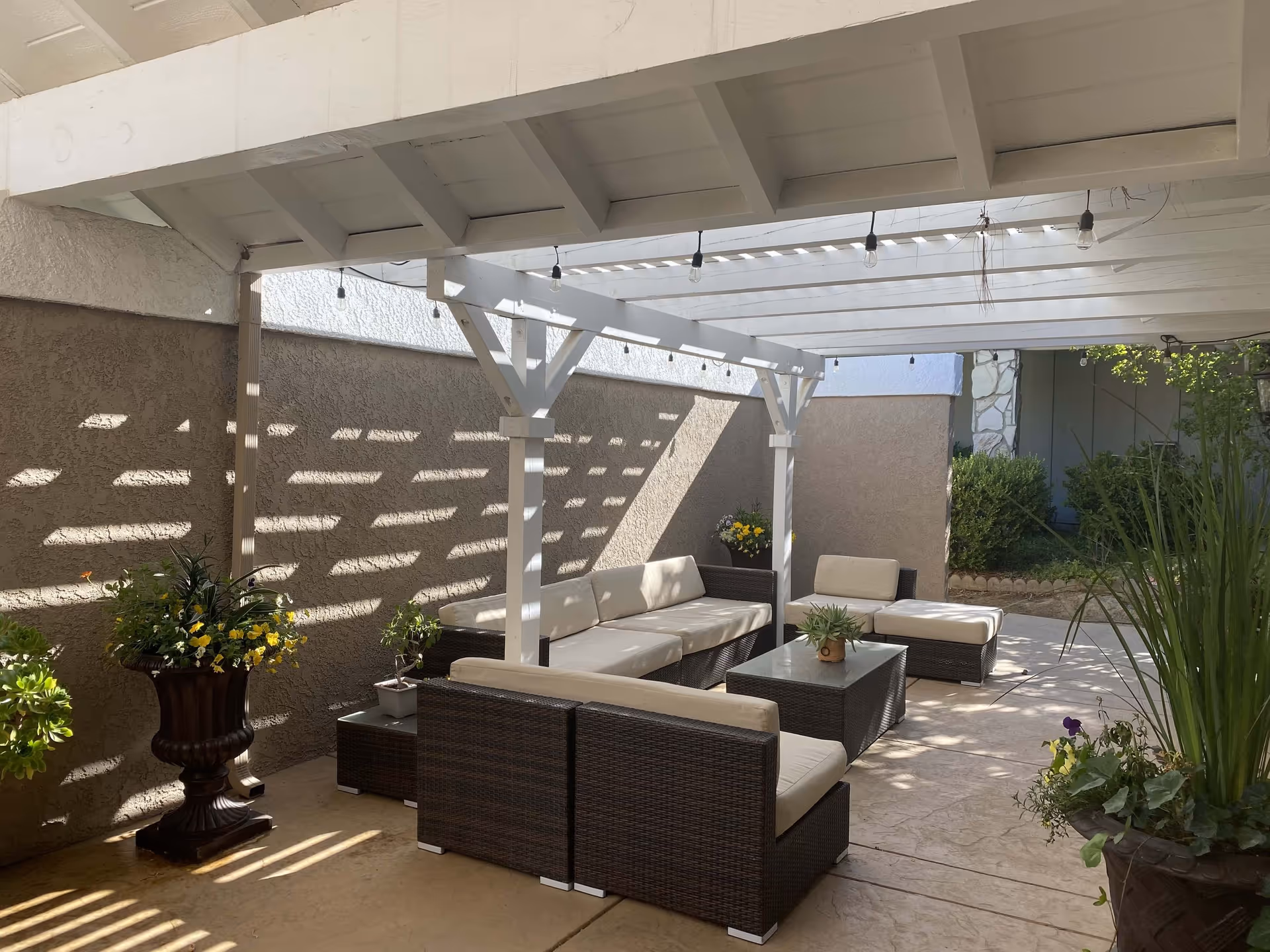 Outdoor patio area with a white pergola overhead casting shadows on the ground and wall. The space is furnished with dark wicker seating including a sofa, chairs, and a coffee table with beige cushions. There are several potted plants and flowers around the seating area, and greenery is visible in the background.