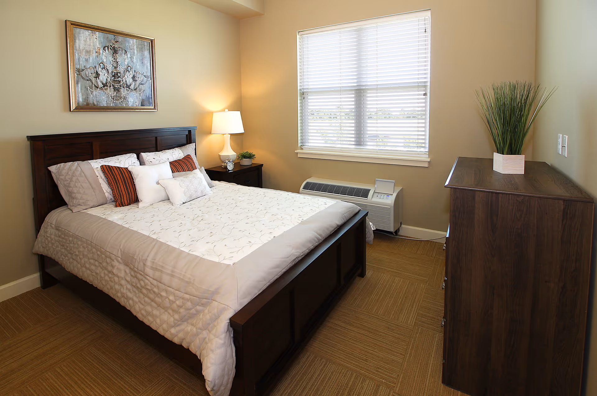 A neatly made bed with a dark wooden frame and multiple pillows in a bedroom. There is a nightstand with a lamp and a small plant beside the bed. A window with blinds lets in natural light, and a wooden dresser with a potted plant on top is positioned against the opposite wall. The walls are painted beige, and a framed artwork hangs above the bed.