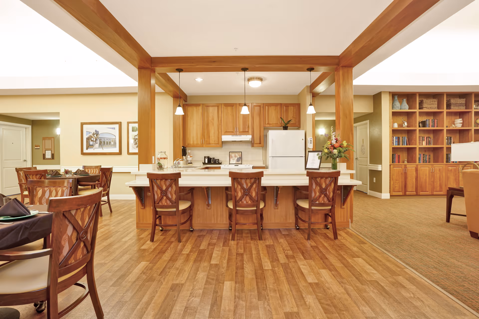 A spacious and well-lit common area featuring a kitchen with wooden cabinets, a white refrigerator, and a large island with four wooden chairs. The room has wooden flooring and is decorated with framed pictures on the wall, a vase with flowers on the counter, and a large wooden bookshelf filled with books and decorative items. Adjacent to the kitchen are dining tables and chairs, and a seating area with armchairs.
