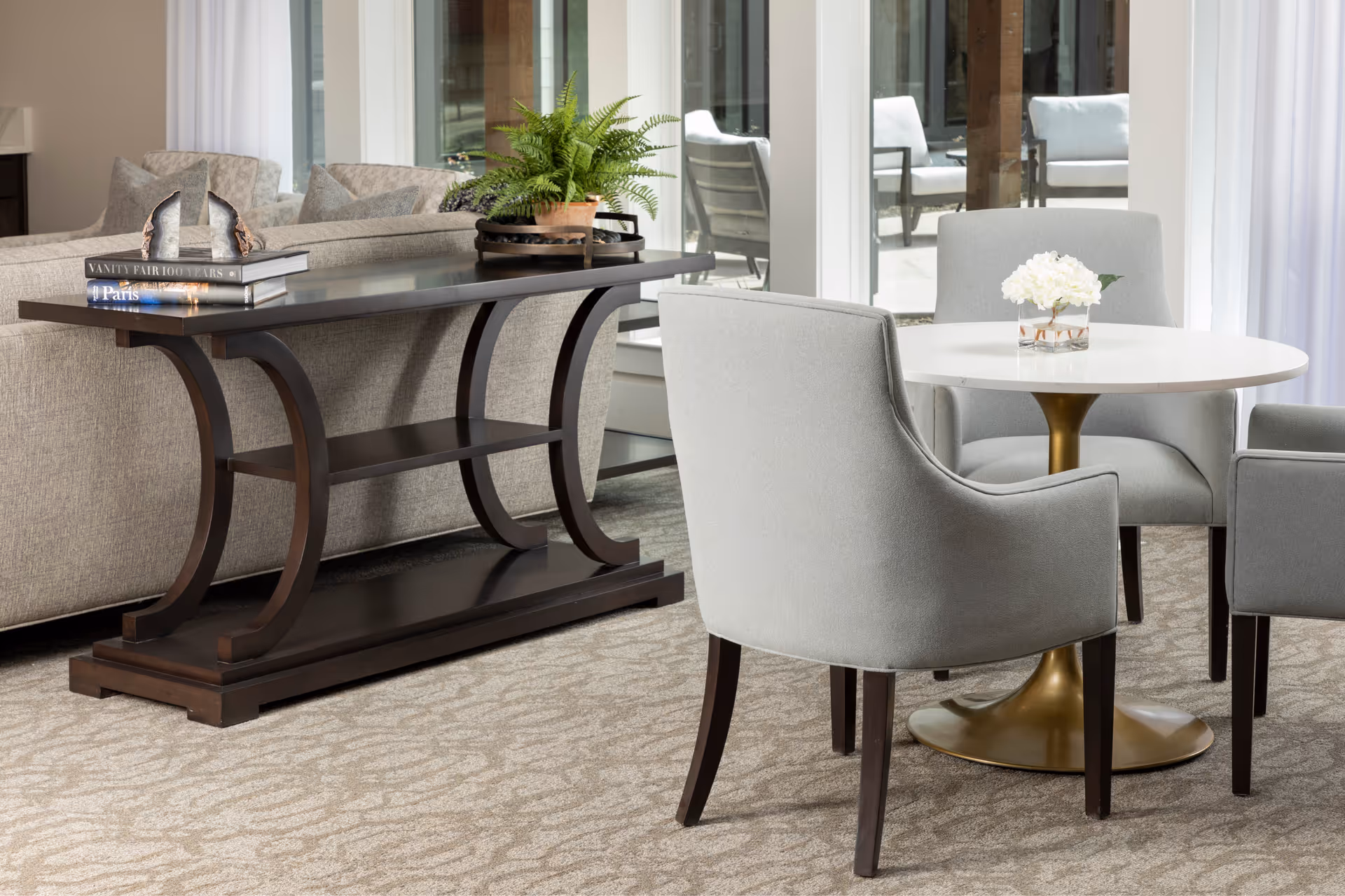A bright, modern living area showing upholstered gray chairs around a round table, a sofa, and a dark wooden console table.