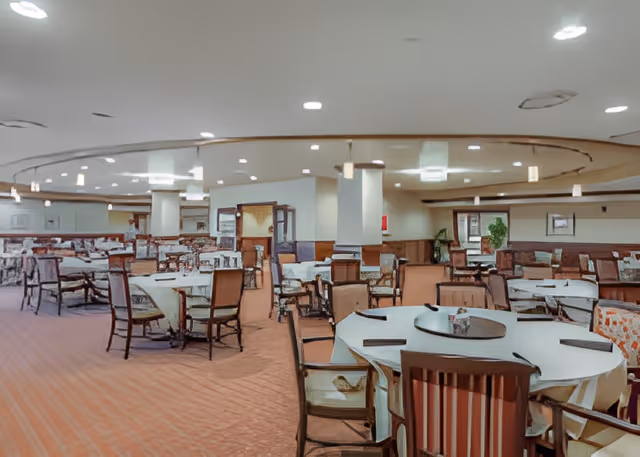 Spacious dining room with multiple round and rectangular tables covered with white tablecloths, surrounded by wooden chairs with cushions. The room has a carpeted floor, recessed ceiling lights, and a circular ceiling design with hanging pendant lights. There are plants and framed pictures on the walls in the background.