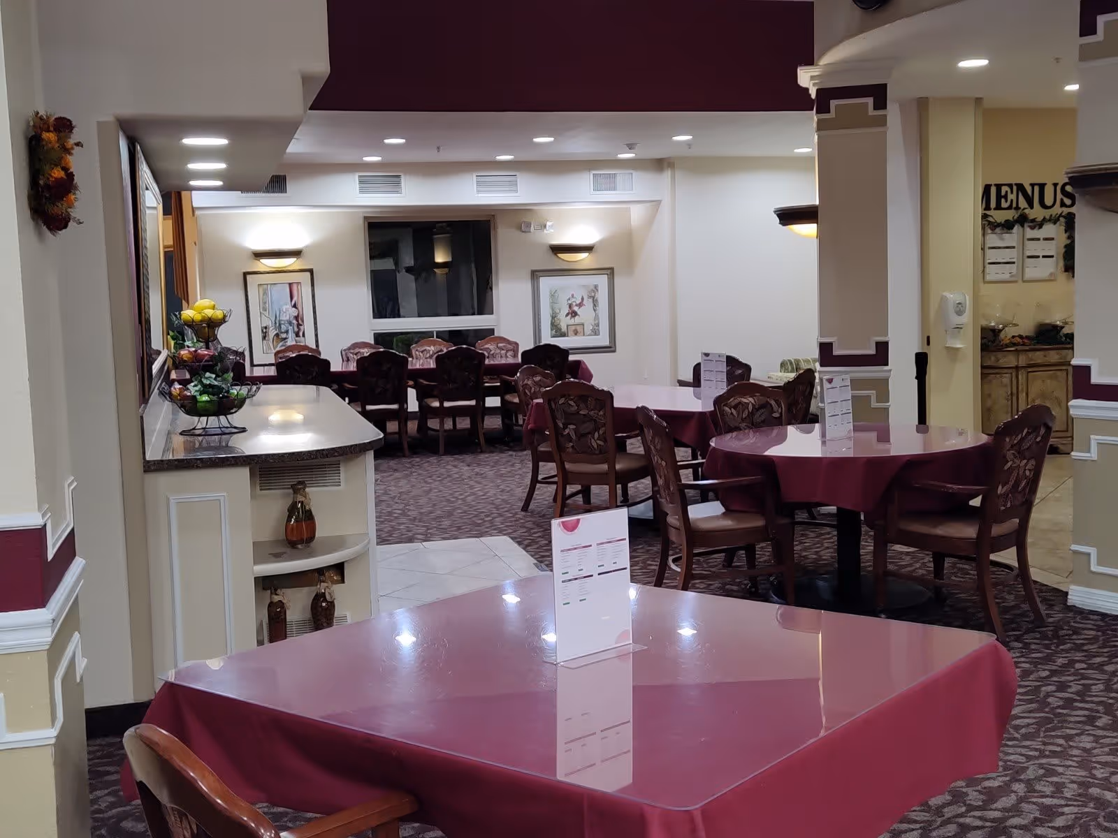 Interior view of a dining area in a senior living facility with multiple tables covered in maroon tablecloths and chairs arranged around them. The room has soft lighting, framed artwork on the walls, and a counter with a fruit basket. A menu board is visible in the background.